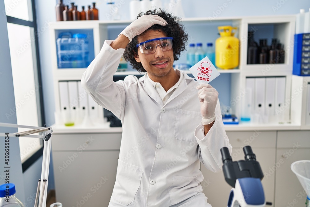 Hispanic man with curly hair working at scientist laboratory holding toxic banner stressed and frustrated with hand on head, surprised and angry face Hispanic man with curly hair working at scientist laboratory holding toxic banner stressed and frustrated with hand on head, surprised and angry face