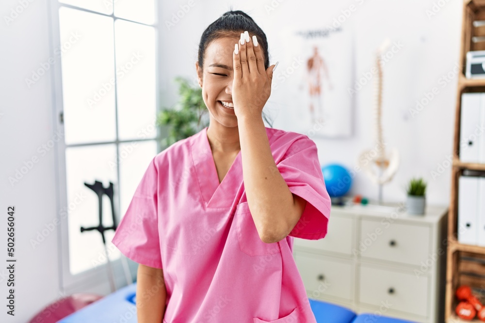 Young hispanic woman working at pain recovery clinic covering one eye with hand, confident smile on face and surprise emotion.