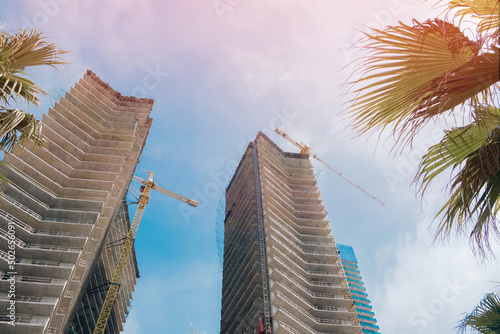 Leaves of a palm tree on a background of high tower crane and residential apartment buildings under construction against blue sky. Real estate development.