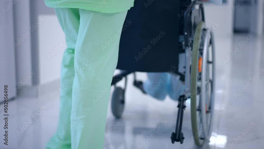 Tracking shot of unrecognizable woman in medical uniform pushing wheelchair with patient in hospital corridor. Young doctor helping girl walking in slow motion. Live camera follows steps Tracking shot of unrecognizable woman in medical uniform pushing wheelchair with patient in hospital corridor. Young doctor helping girl walking in slow motion. Live camera follows steps