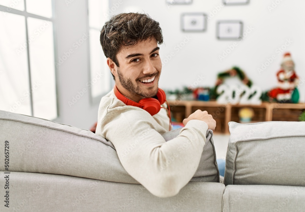 Young hispanic man celebrating christmas using headphones sitting on the sofa at home.