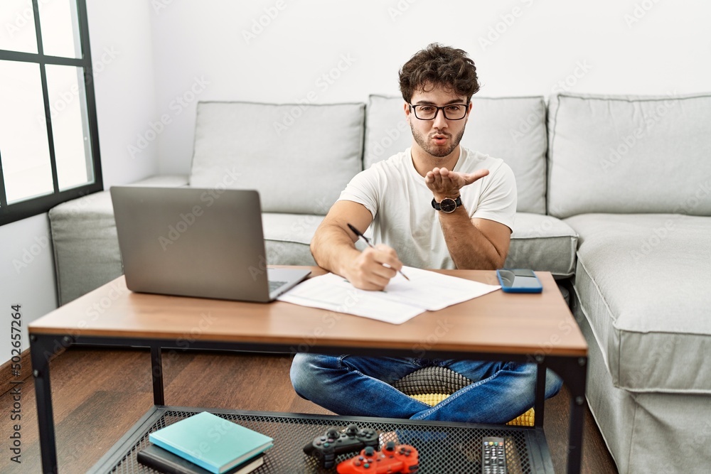Hispanic man doing papers at home looking at the camera blowing a kiss with hand on air being lovely and sexy. love expression. Hispanic man doing papers at home looking at the camera blowing a kiss with hand on air being lovely and sexy. love expression.