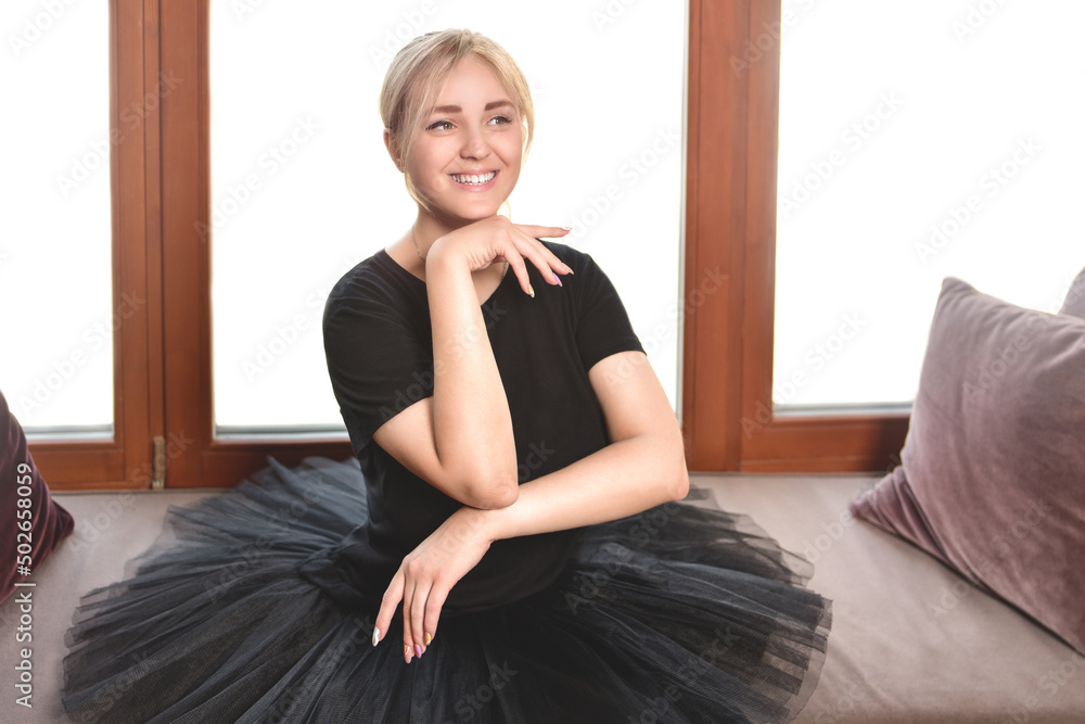 smiling ballerina in a black tutu sits in front of the window with her chin tucked in smiling ballerina in a black tutu sits in front of the window with her chin tucked in