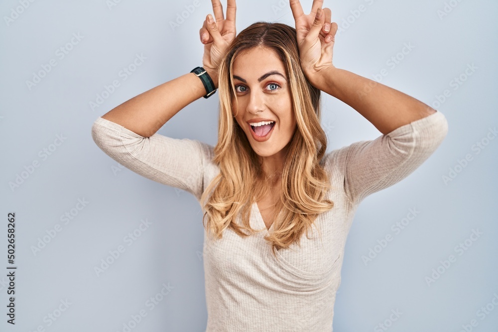 Young blonde woman standing over isolated background posing funny and crazy with fingers on head as bunny ears, smiling cheerful
