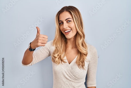 Young blonde woman standing over isolated background doing happy thumbs up gesture with hand. approving expression looking at the camera showing success.