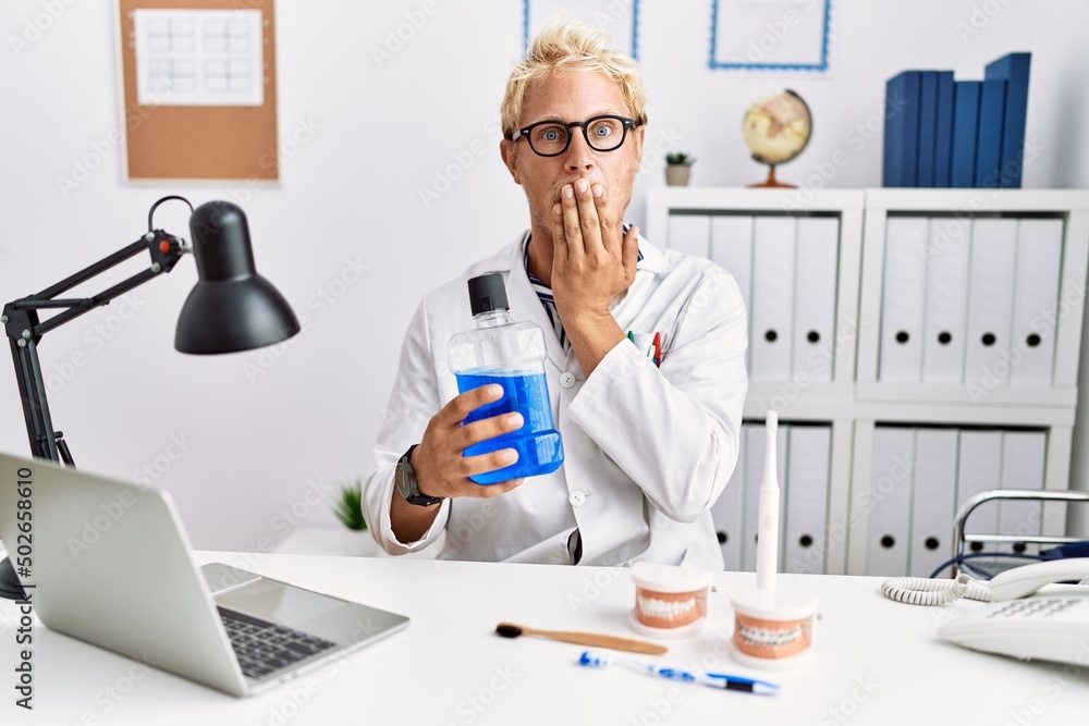 Young blond dentist man holding mouthwash at clinic covering mouth with hand, shocked and afraid for mistake. surprised expression Young blond dentist man holding mouthwash at clinic covering mouth with hand, shocked and afraid for mistake. surprised expression
