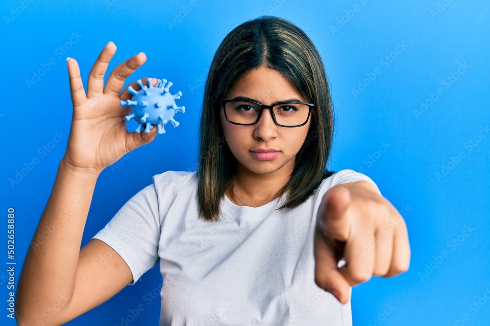 Young latin woman holding virus toy pointing with finger to the camera and to you, confident gesture looking serious Young latin woman holding virus toy pointing with finger to the camera and to you, confident gesture looking serious