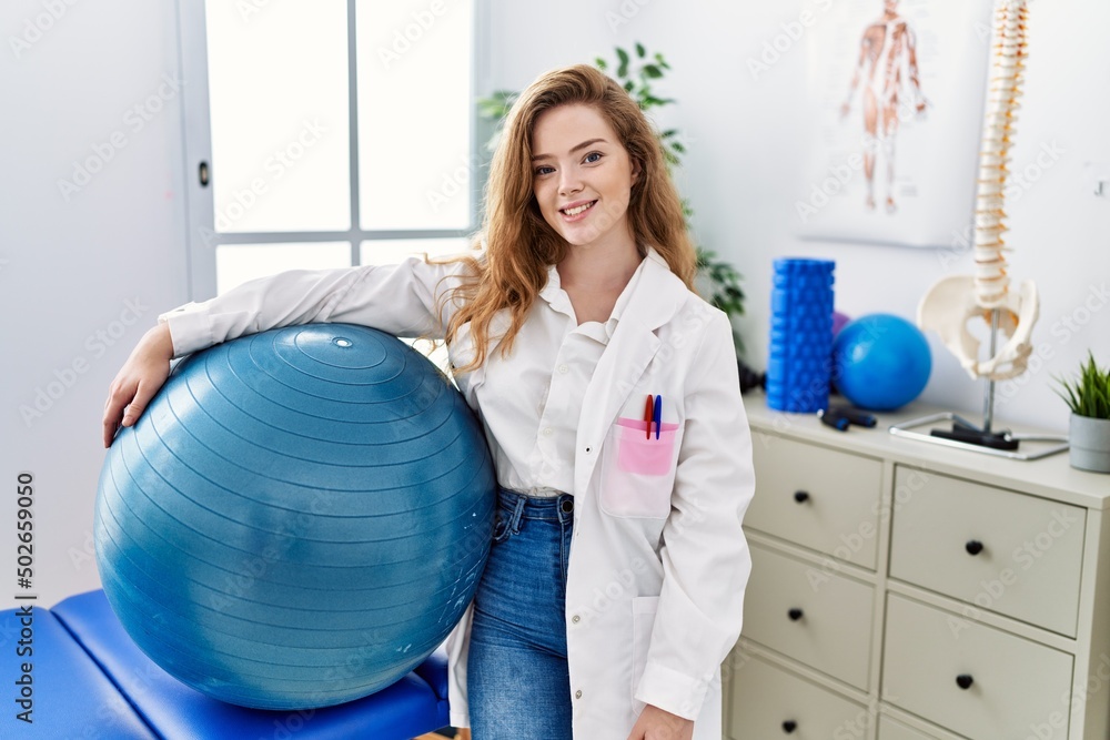 Fototapeta premium Young caucasian woman working at rehabilitation clinic holding pilates ball looking positive and happy standing and smiling with a confident smile showing teeth