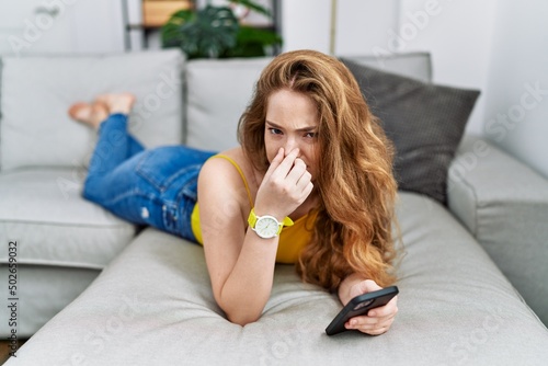 Young caucasian woman lying on the sofa using smartphone smelling something stinky and disgusting, intolerable smell, holding breath with fingers on nose. bad smell photo