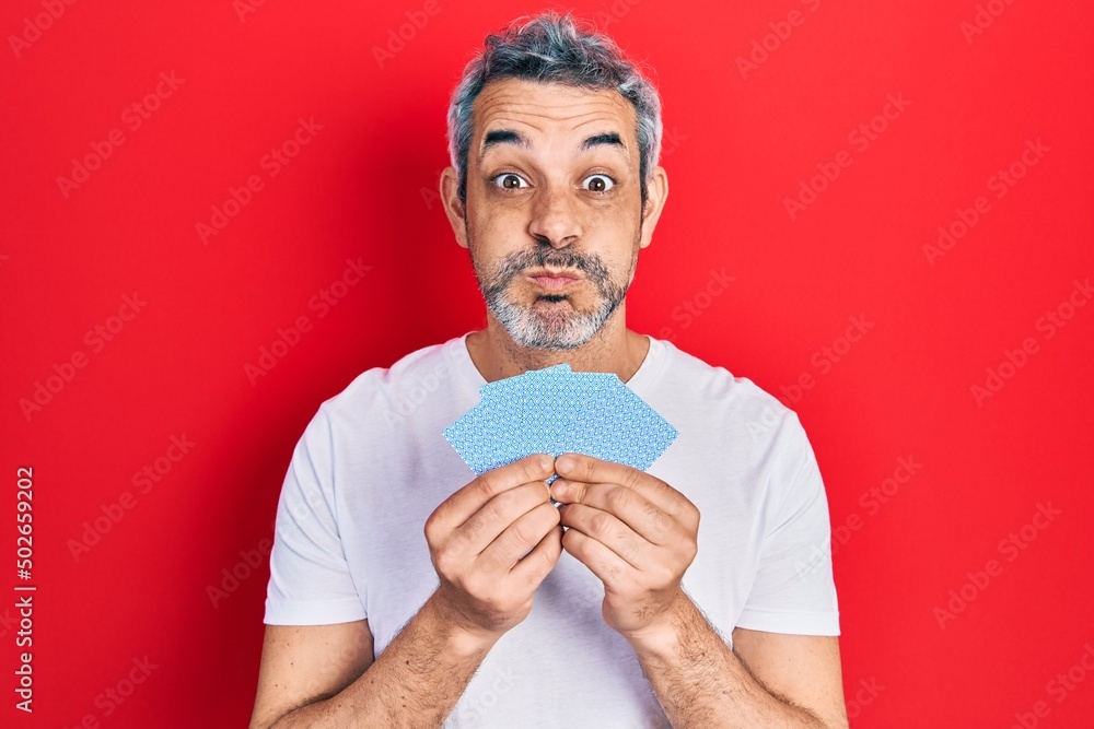 Handsome middle age man with grey hair covering face with cards puffing cheeks with funny face. mouth inflated with air, catching air. Handsome middle age man with grey hair covering face with cards puffing cheeks with funny face. mouth inflated with air, catching air.
