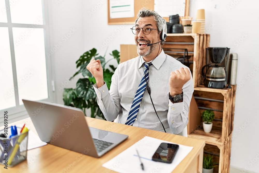 Middle age hispanic business man working at the office wearing operator headset celebrating surprised and amazed for success with arms raised and open eyes. winner concept. Middle age hispanic business man working at the office wearing operator headset celebrating surprised and amazed for success with arms raised and open eyes. winner concept.