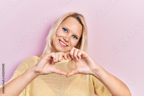 Beautiful caucasian blonde woman wearing casual winter sweater smiling in love doing heart symbol shape with hands. romantic concept.