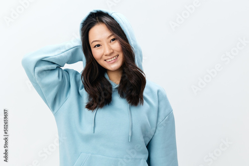 Fashionable smiling happy cute Asian young female in blue sweatshirt puts on the hood posing isolated on over white studio background. The best offer for ad. Fashion New Collection Ad concept