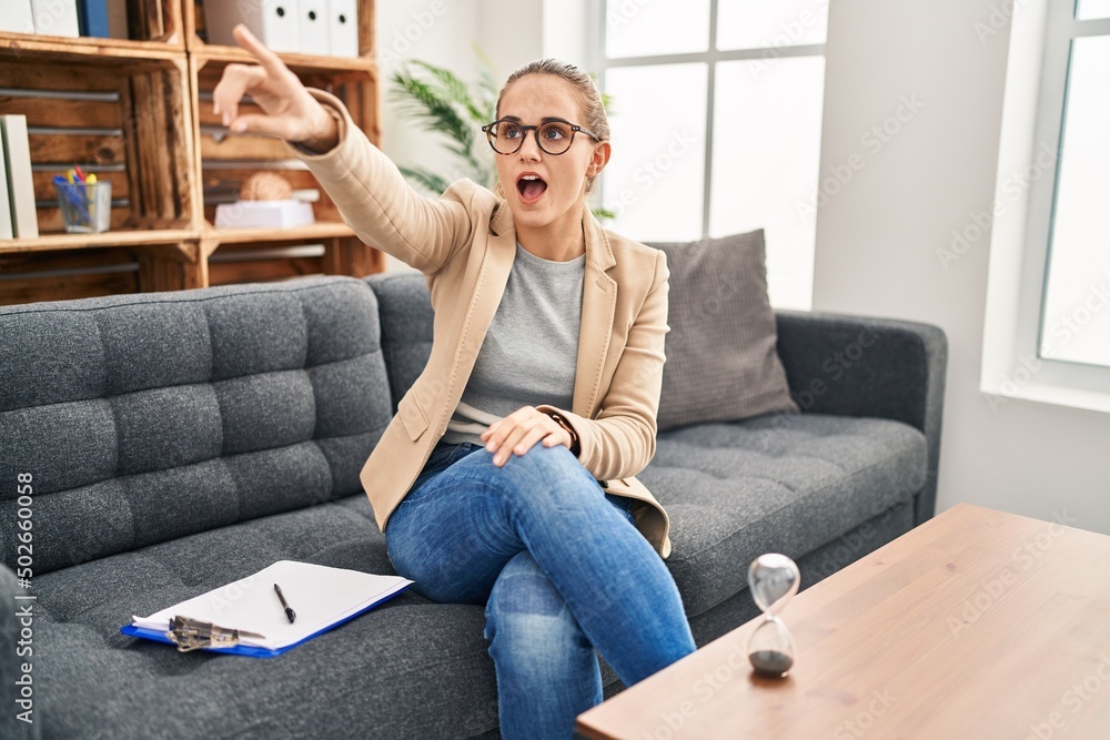 Young woman working at consultation office pointing with finger surprised ahead, open mouth amazed expression, something on the front Young woman working at consultation office pointing with finger surprised ahead, open mouth amazed expression, something on the front