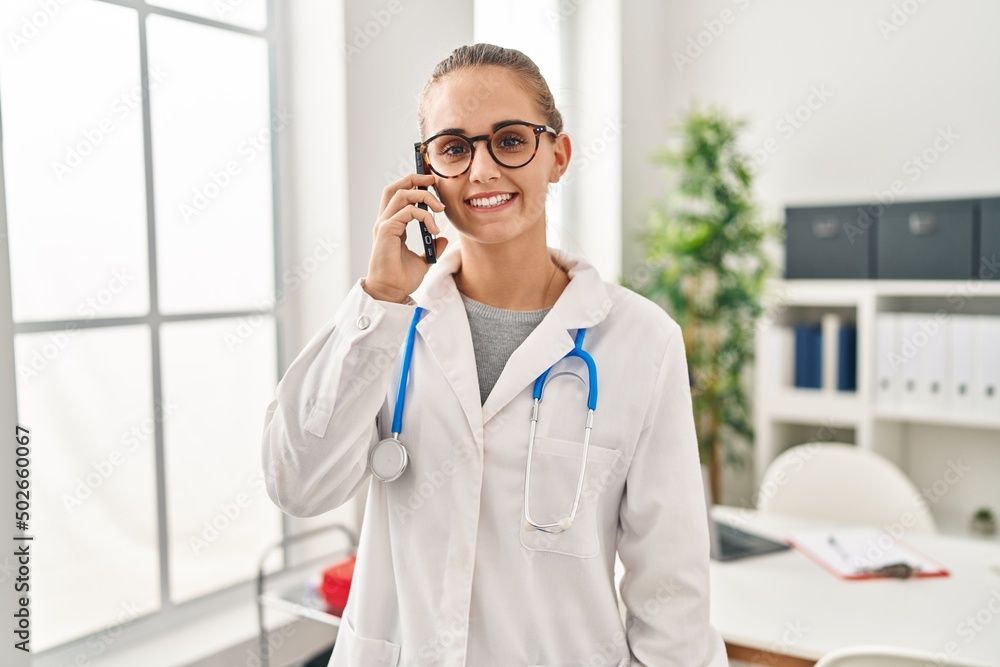 Young blonde woman wearing doctor uniform talking on the smartphone working at clinic Young blonde woman wearing doctor uniform talking on the smartphone working at clinic