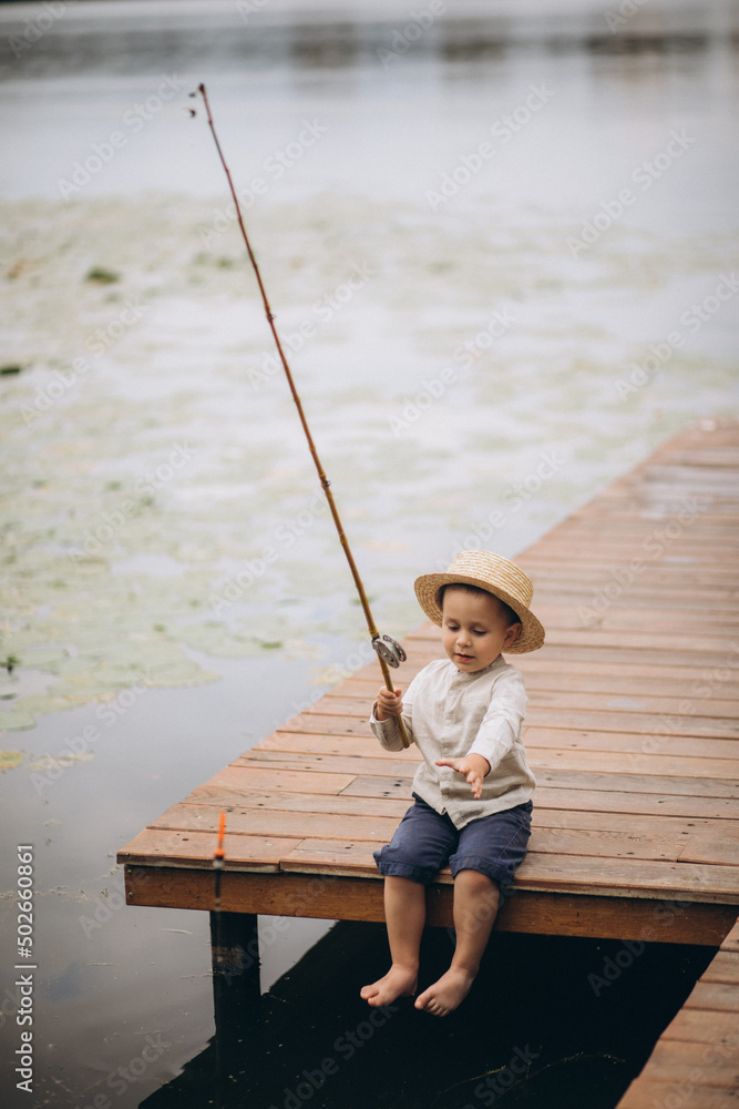 Little boy is fishing on the pier wearing a hat and a fishing rod in his hands Little boy is fishing on the pier wearing a hat and a fishing rod in his hands