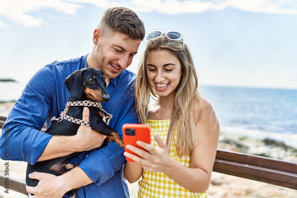 Young caucasian couple using smartphone standing with dog at the beach.