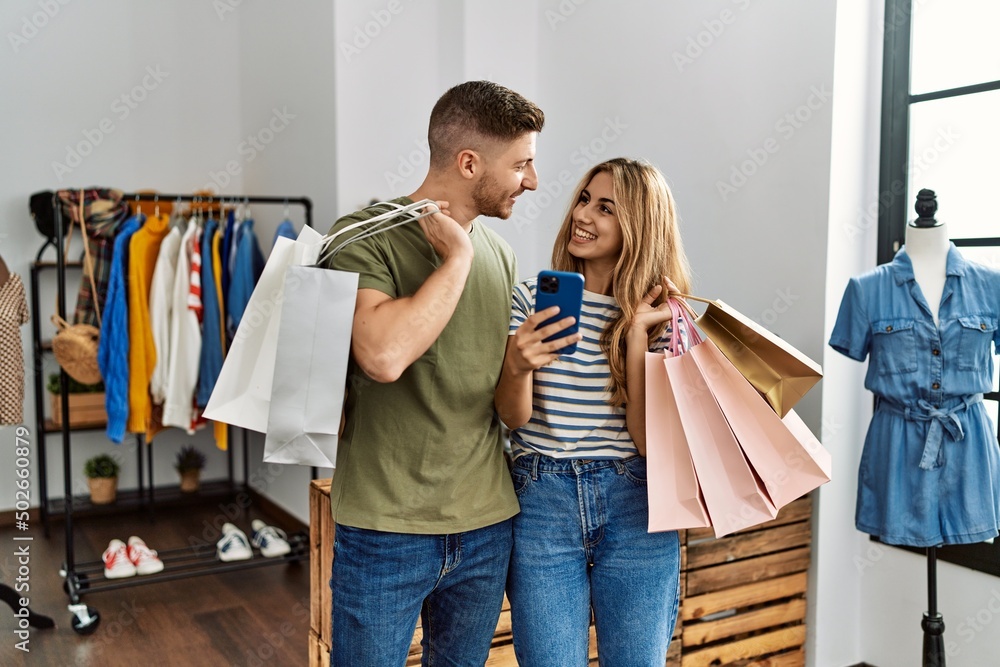 Young hispanic customer couple holding shopping bags and using smartphone at clothing store. Young hispanic customer couple holding shopping bags and using smartphone at clothing store.