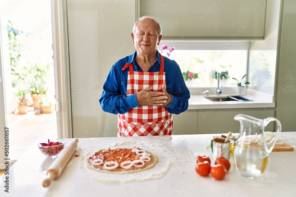 Senior man with grey hair cooking pizza at home kitchen smiling with hands on chest with closed eyes and grateful gesture on face. health concept. Senior man with grey hair cooking pizza at home kitchen smiling with hands on chest with closed eyes and grateful gesture on face. health concept.