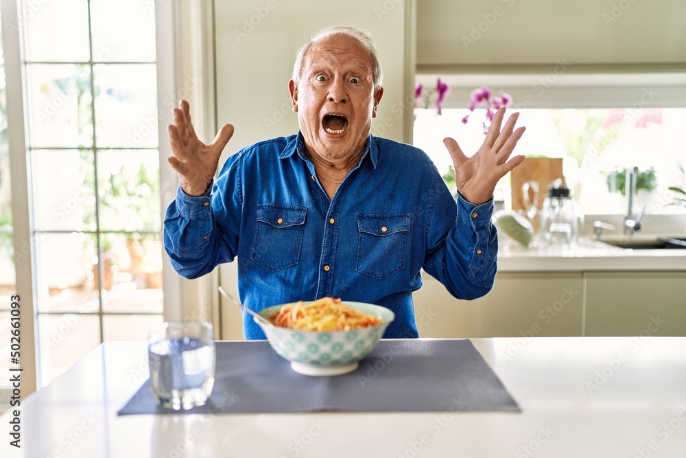 Senior man with grey hair eating pasta spaghetti at home celebrating crazy and amazed for success with arms raised and open eyes screaming excited. winner concept Senior man with grey hair eating pasta spaghetti at home celebrating crazy and amazed for success with arms raised and open eyes screaming excited. winner concept