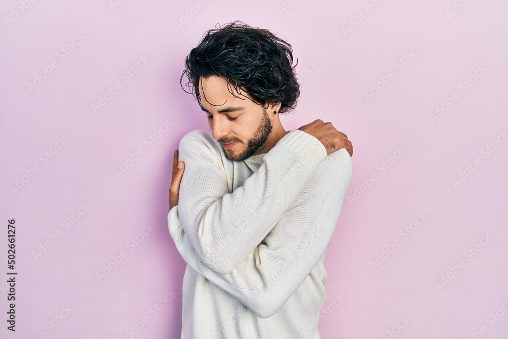 Handsome hispanic man wearing casual white sweater hugging oneself happy and positive, smiling confident. self love and self care Handsome hispanic man wearing casual white sweater hugging oneself happy and positive, smiling confident. self love and self care