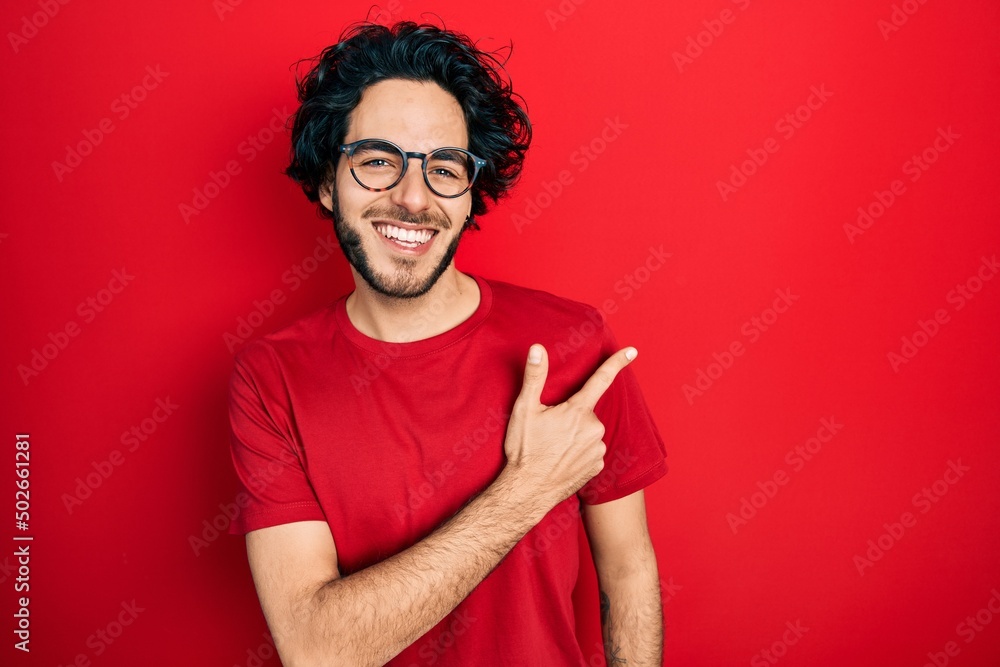 Handsome hispanic man wearing casual t shirt and glasses cheerful with a smile of face pointing with hand and finger up to the side with happy and natural expression on face Handsome hispanic man wearing casual t shirt and glasses cheerful with a smile of face pointing with hand and finger up to the side with happy and natural expression on face