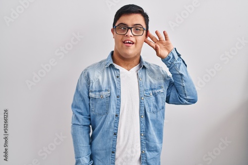 Young hispanic man with down syndrome wearing casual denim jacket over white background smiling with hand over ear listening and hearing to rumor or gossip. deafness concept.