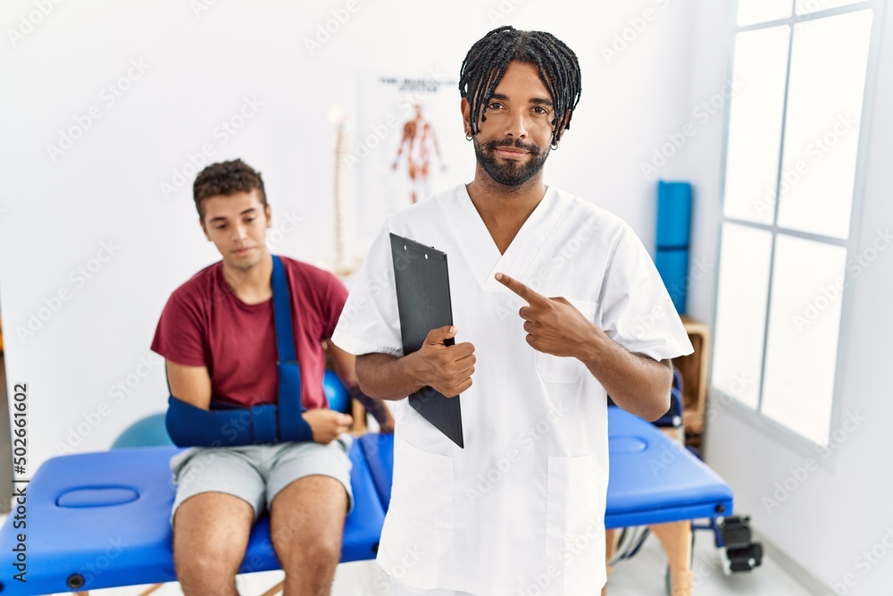 Young hispanic man working at pain recovery clinic with a man with broken arm pointing with hand finger to the side showing advertisement, serious and calm face Young hispanic man working at pain recovery clinic with a man with broken arm pointing with hand finger to the side showing advertisement, serious and calm face