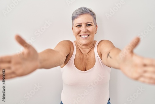 Middle age caucasian woman standing over white background looking at the camera smiling with open arms for hug. cheerful expression embracing happiness.