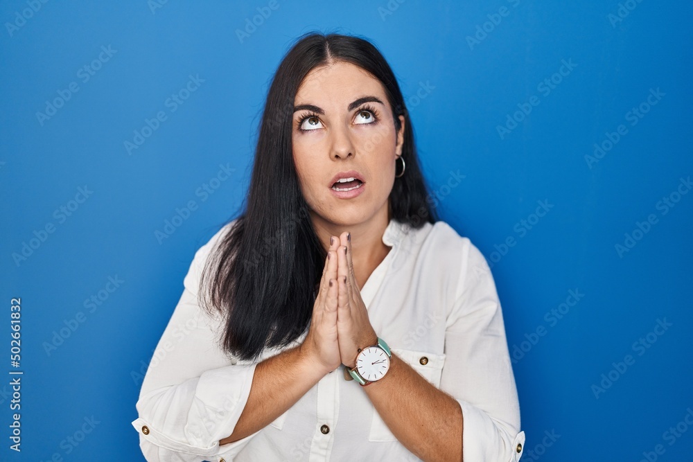 Young hispanic woman standing over blue background begging and praying with hands together with hope expression on face very emotional and worried. begging. Young hispanic woman standing over blue background begging and praying with hands together with hope expression on face very emotional and worried. begging.
