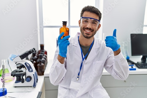 Young hispanic man working at scientist laboratory holding bottle smiling happy and positive, thumb up doing excellent and approval sign