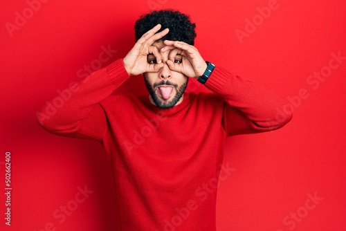 Young arab man with beard wearing casual red sweater doing ok gesture like binoculars sticking tongue out, eyes looking through fingers. crazy expression.