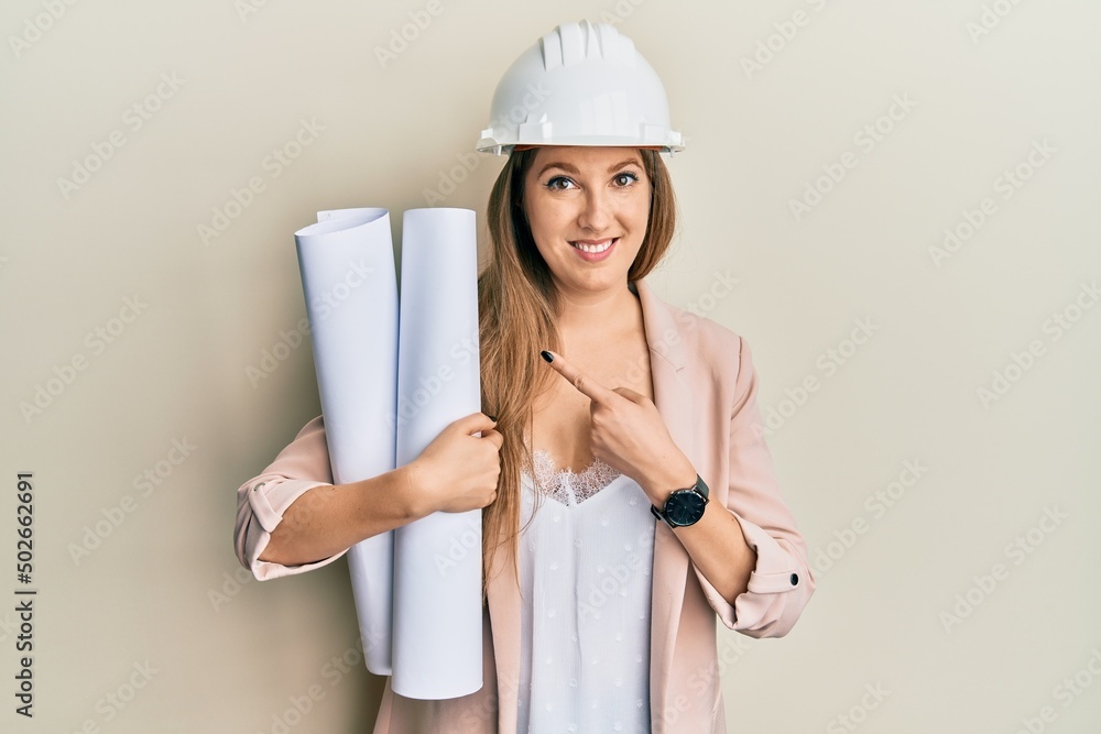 Young blonde woman wearing safety helmet holding blueprints smiling happy pointing with hand and finger Young blonde woman wearing safety helmet holding blueprints smiling happy pointing with hand and finger