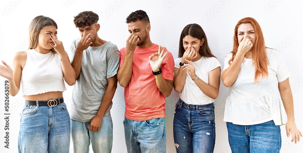 Group of young friends standing together over isolated background smelling something stinky and disgusting, intolerable smell, holding breath with fingers on nose. bad smell - obrazy, fototapety, plakaty 