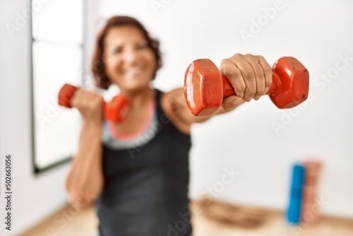 Middle age hispanic sporty woman smiling happy training using dumbbells at sport center.