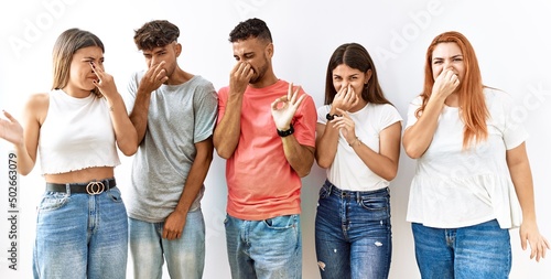 Bild auf Leinwand Group of young friends standing together over isolated background smelling something stinky and disgusting, intolerable smell, holding breath with fingers on nose