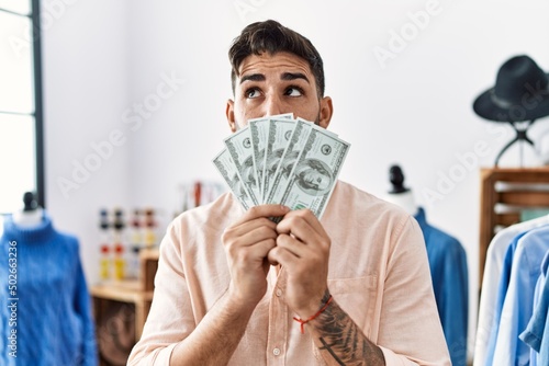 Young hispanic man holding dollars banknotes at retail shop smiling looking to the side and staring away thinking.