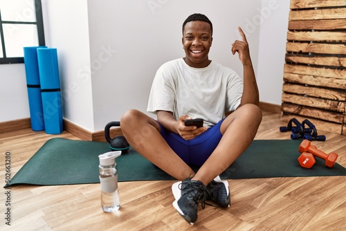 Young african man sitting on training mat at the gym using smartphone smiling amazed and surprised and pointing up with fingers and raised arms.