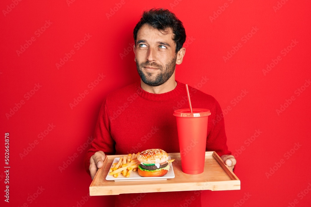 Handsome man with beard eating a tasty classic burger with fries and soda smiling looking to the side and staring away thinking. Handsome man with beard eating a tasty classic burger with fries and soda smiling looking to the side and staring away thinking.