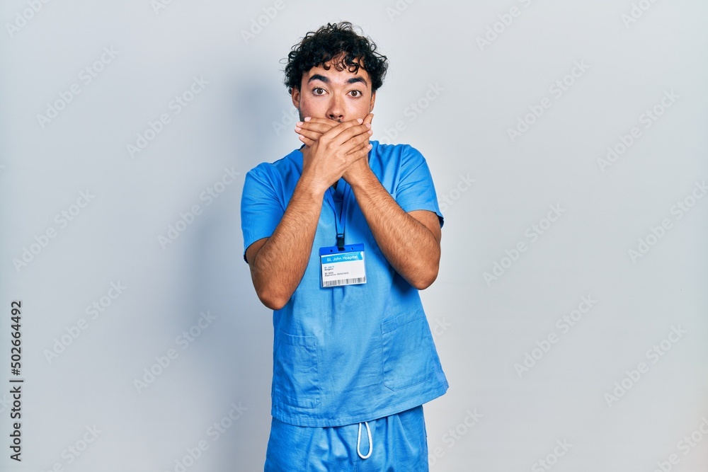 Young hispanic man wearing blue male nurse uniform shocked covering mouth with hands for mistake. secret concept. Young hispanic man wearing blue male nurse uniform shocked covering mouth with hands for mistake. secret concept.