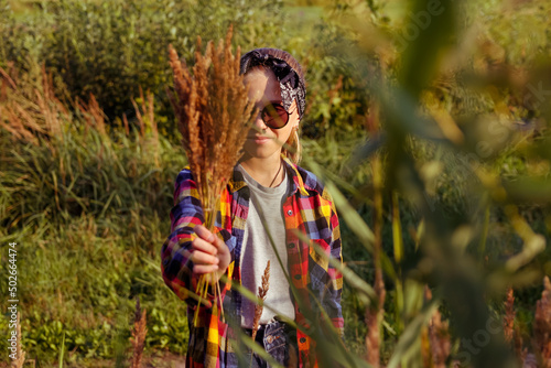 Defocus teen or preteen girl walking on nature background. Little kid girl showing bunch of pampas grass. Blurred reed on foreground. Hipster generation z Bandana  sunglasses. Summertime Out of focus