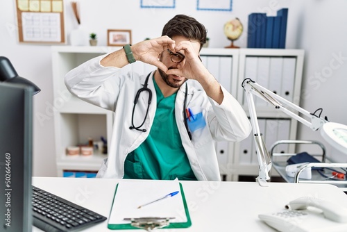 Young man with beard wearing doctor uniform and stethoscope at the clinic doing heart shape with hand and fingers smiling looking through sign