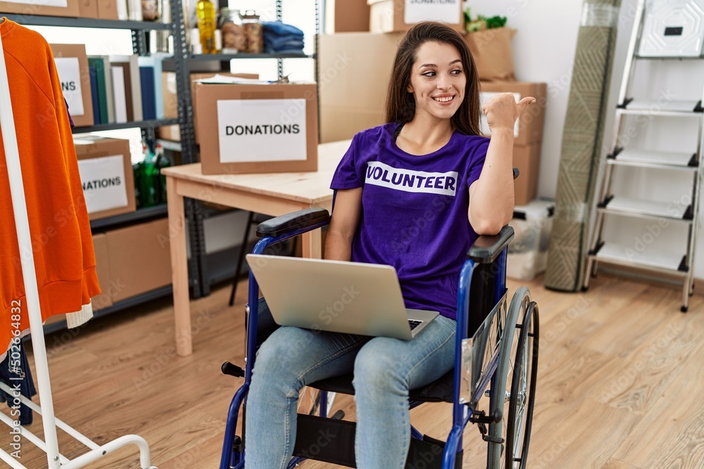 Young brunette woman as volunteer on donations stand sitting on wheelchair pointing thumb up to the side smiling happy with open mouth Young brunette woman as volunteer on donations stand sitting on wheelchair pointing thumb up to the side smiling happy with open mouth