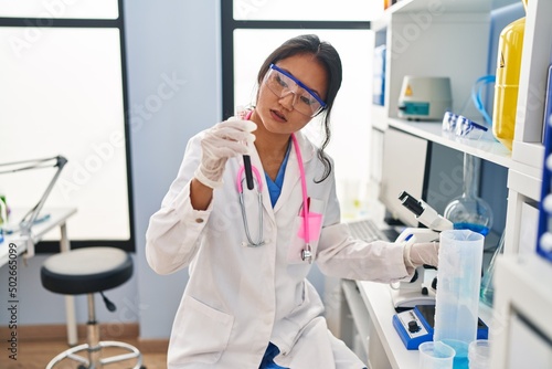 Young chinese woman wearing scientist uniform holding blood test tube at laboratory