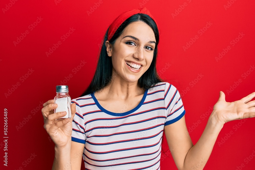 Young hispanic woman holding salt shaker celebrating achievement with happy smile and winner expression with raised hand Young hispanic woman holding salt shaker celebrating achievement with happy smile and winner expression with raised hand