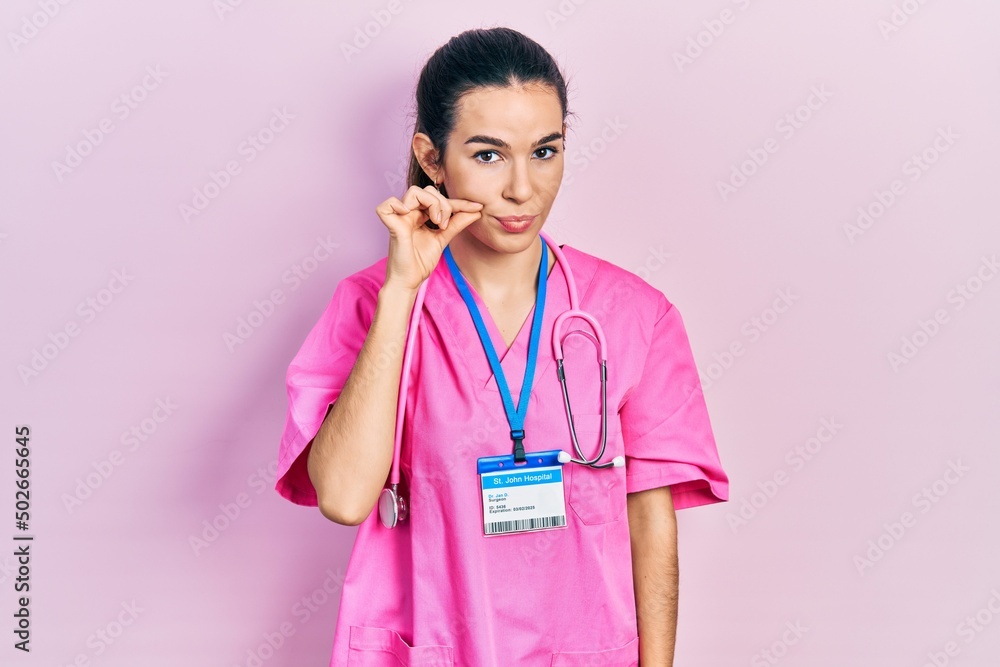 Young brunette woman wearing doctor uniform and stethoscope mouth and lips shut as zip with fingers. secret and silent, taboo talking Young brunette woman wearing doctor uniform and stethoscope mouth and lips shut as zip with fingers. secret and silent, taboo talking