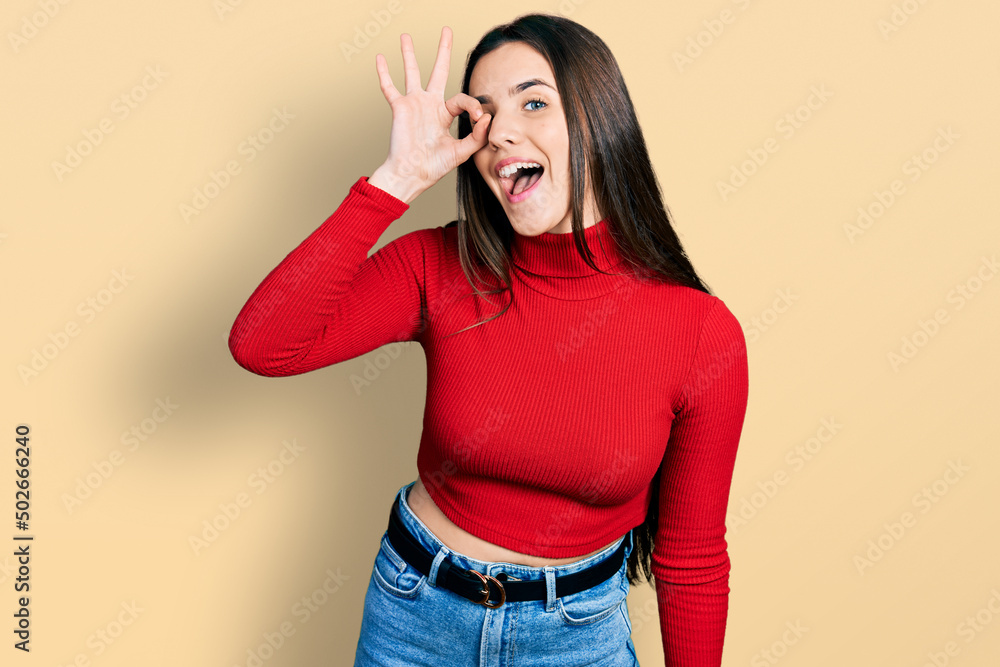 Young brunette teenager wearing red turtleneck sweater doing ok gesture with hand smiling, eye looking through fingers with happy face. Young brunette teenager wearing red turtleneck sweater doing ok gesture with hand smiling, eye looking through fingers with happy face.