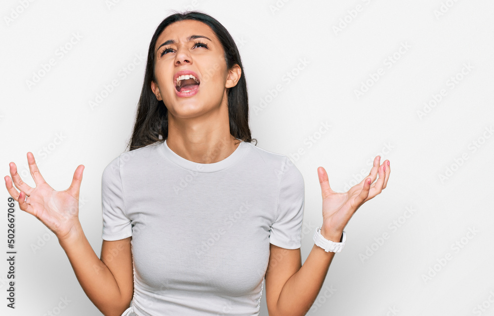 Young hispanic girl wearing casual white t shirt crazy and mad shouting and yelling with aggressive expression and arms raised. frustration concept. Young hispanic girl wearing casual white t shirt crazy and mad shouting and yelling with aggressive expression and arms raised. frustration concept.