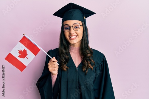 Young hispanic woman wearing graduation uniform holding canada flag looking positive and happy standing and smiling with a confident smile showing teeth