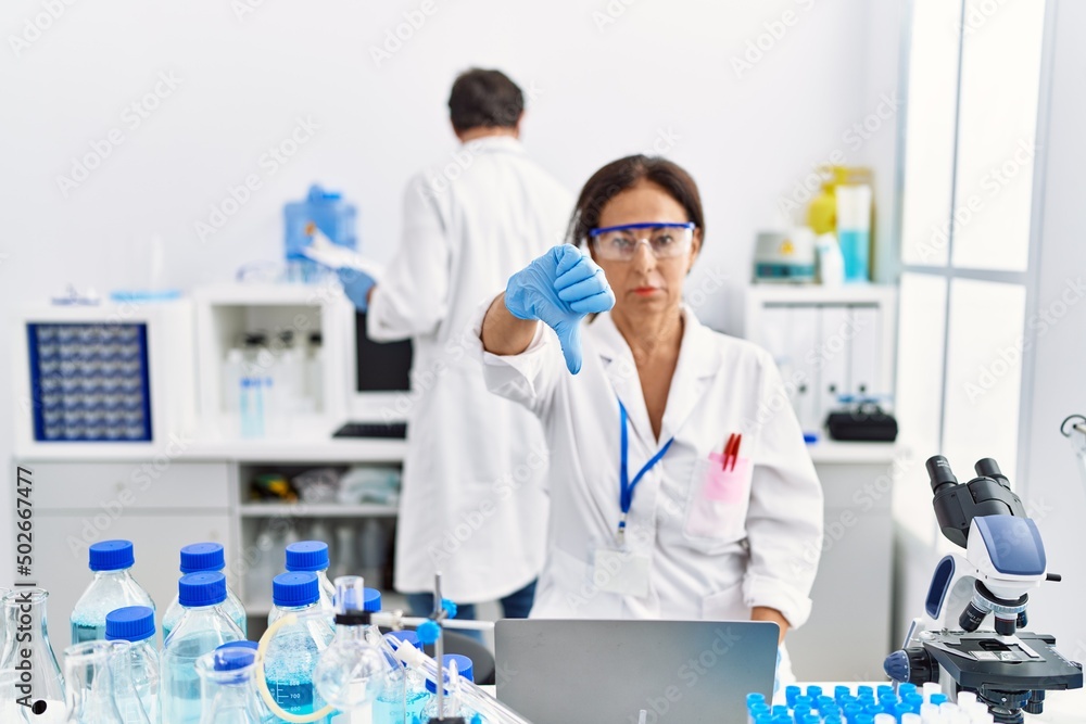 Middle age woman working at scientist laboratory with angry face, negative sign showing dislike with thumbs down, rejection concept Middle age woman working at scientist laboratory with angry face, negative sign showing dislike with thumbs down, rejection concept